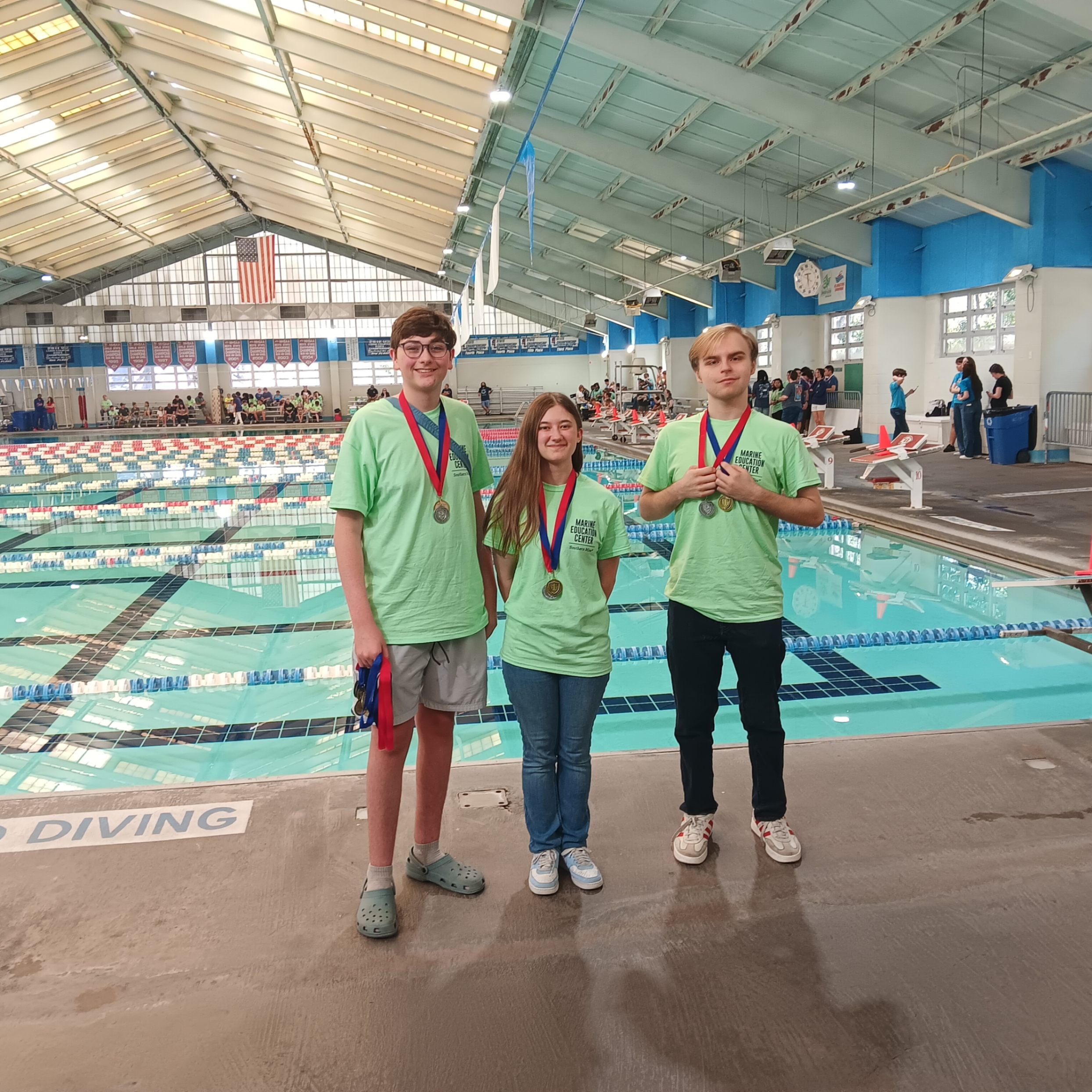 Three students stand on the deck of an indoor swimming facility wearing matching light green “Murray County Schools Academic Team” T‑shirts. Each student has a medal around their neck, smiling toward the camera. Behind them is a large competition pool with lane dividers, diving platforms, and an American flag hanging at the far end. Spectators and participants are visible in the background.  If you want a shorter or 